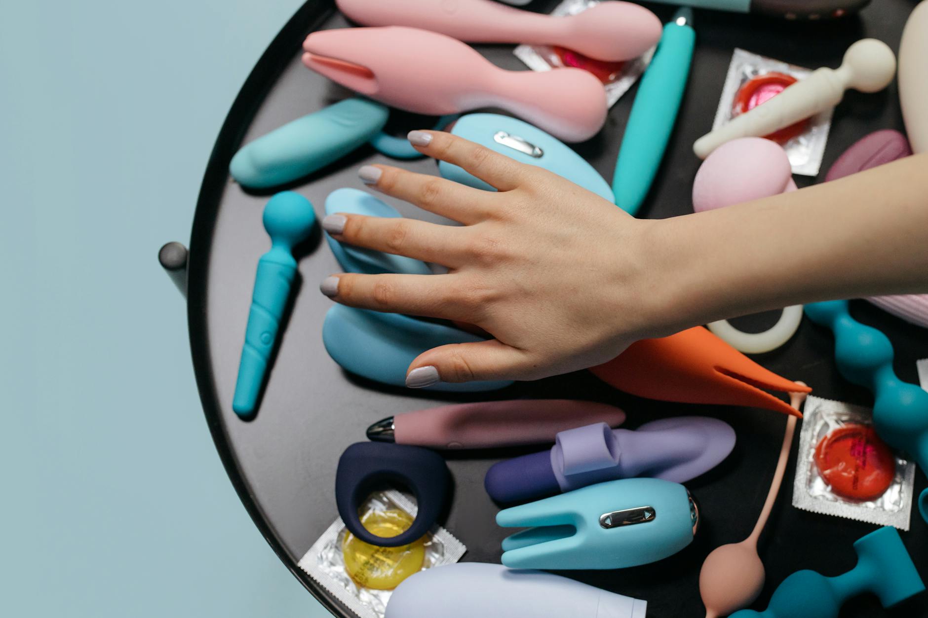 A hand reaching over a variety of colorful sex toys on a table, showing options for couples play.
