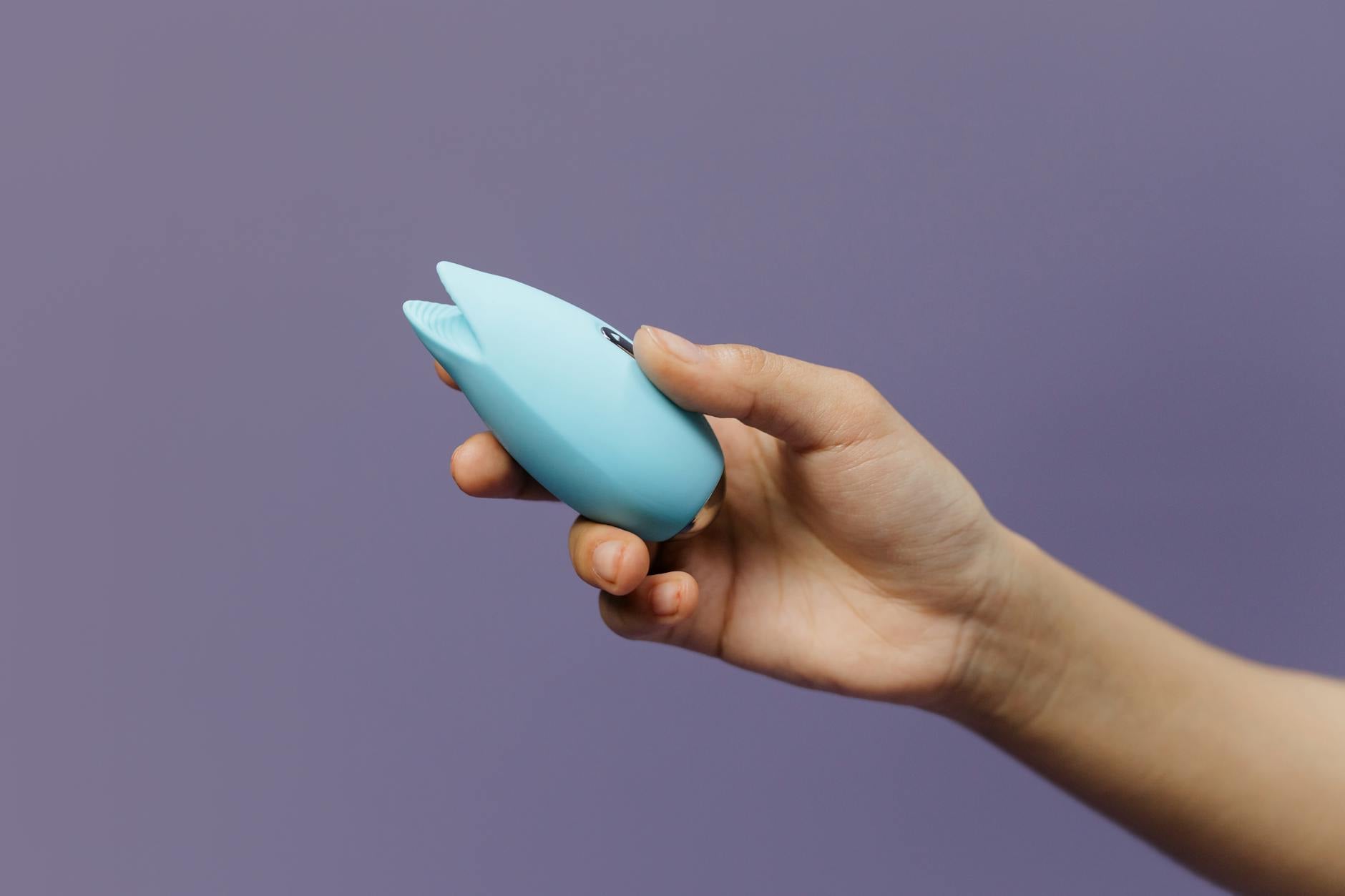 A hand holding a blue silicone vibrator against a solid purple background